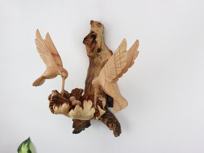 May include: A wooden sculpture of two hummingbirds perched on a branch with a nest of eggs. The birds are carved in detail and have a natural wood finish.