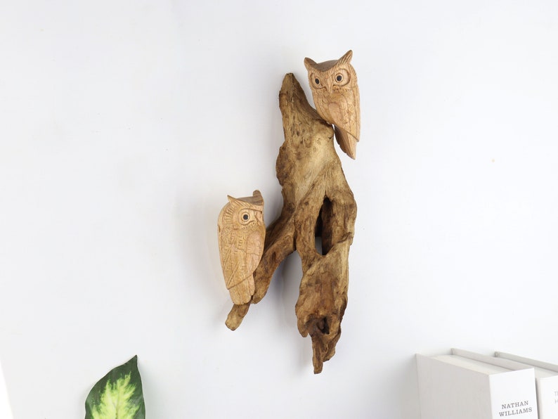 May include: Two intricately carved wooden owls are perched on a piece of driftwood. The owls are facing forward with their wings outstretched. The driftwood is a light brown colour and has a natural, rough texture.