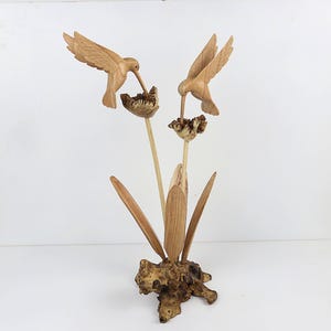 May include: A wooden sculpture of two hummingbirds perched on a flower with leaves. The sculpture is made of light brown wood and has a natural finish.