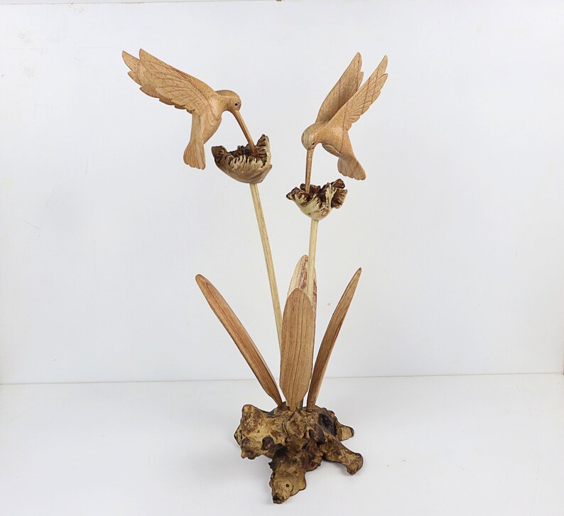 May include: A wooden sculpture of two hummingbirds perched on a flower with leaves. The sculpture is made of light brown wood and has a natural finish.