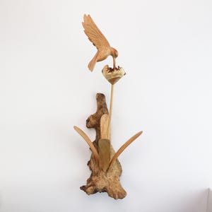 May include: A wooden sculpture of a hummingbird perched on a flower, with its beak in the flower. The sculpture is made of driftwood and has a natural, rustic look.