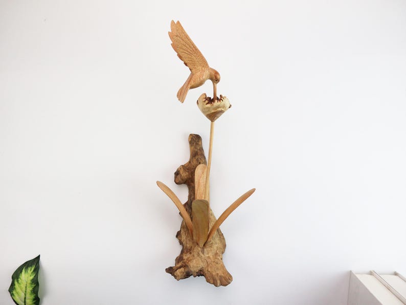 May include: A wooden sculpture of a hummingbird perched on a flower, with its beak in the flower. The sculpture is made of driftwood and has a natural, rustic look.