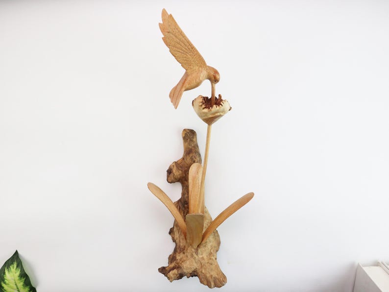 May include: A wooden sculpture of a hummingbird perched on a flower. The sculpture is made of natural wood and has a rustic, hand-crafted look.