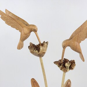 May include: Two wooden hummingbirds carved in detail, perched on a wooden flower stem with brown and white flowers. The hummingbirds are in flight, with their beaks extended as if feeding from the flowers.