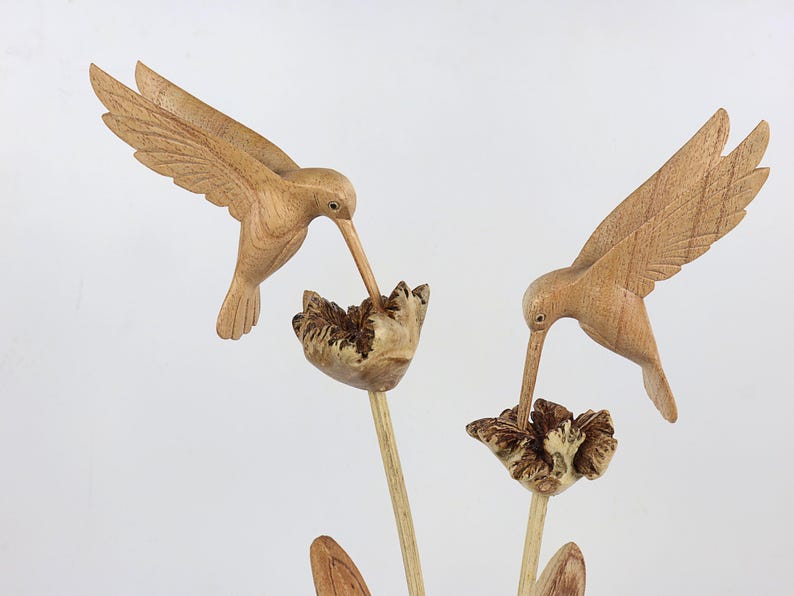 May include: Two wooden hummingbirds carved in detail, perched on a wooden flower stem with brown and white flowers. The hummingbirds are in flight, with their beaks extended as if feeding from the flowers.