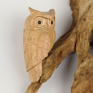 May include: A wooden owl sculpture perched on a branch of driftwood. The owl is carved with intricate details, including its eyes, feathers, and beak. The driftwood is a natural brown colour with a rough texture.