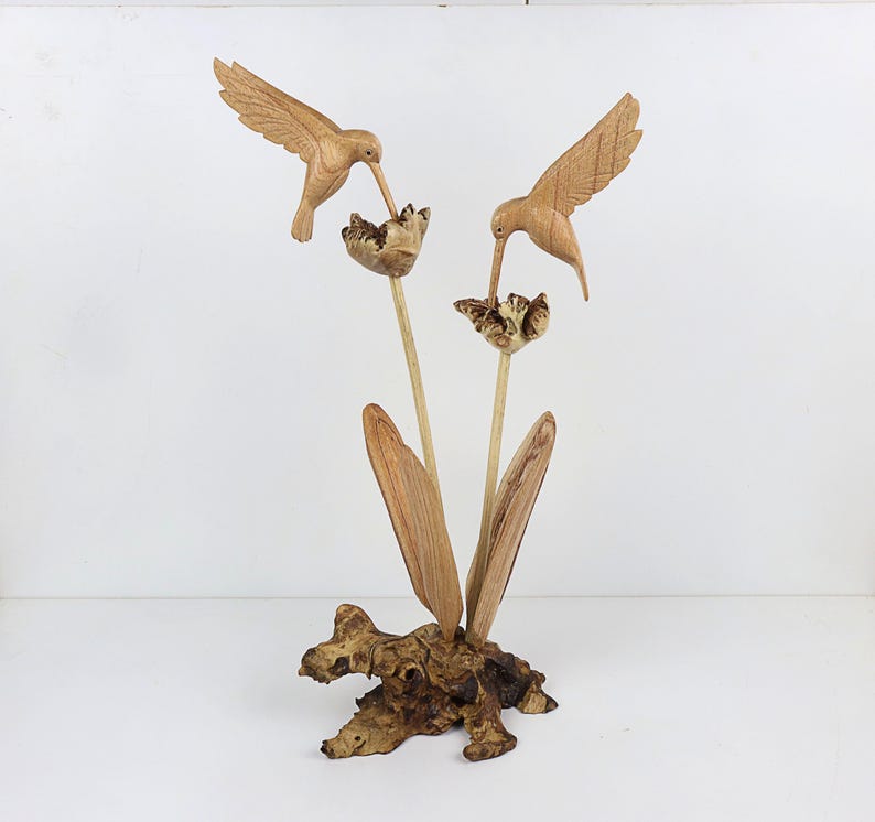 May include: A wooden sculpture of two hummingbirds perched on a branch with flowers. The birds are carved in detail and the flowers are made of wood.
