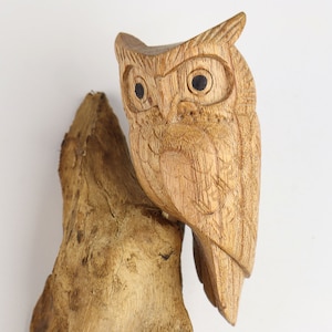 May include: A wooden owl sculpture perched on a piece of driftwood. The owl has detailed carvings and black eyes.
