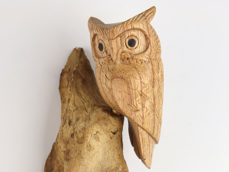 May include: A wooden owl sculpture perched on a piece of driftwood. The owl has detailed carvings and black eyes.