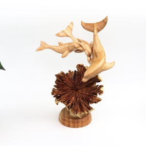 May include: A wooden sculpture of three dolphins leaping out of a coral reef. The dolphins are carved in a realistic style and are painted in shades of brown. The coral reef is carved in a more abstract style and is painted in shades of brown and red.