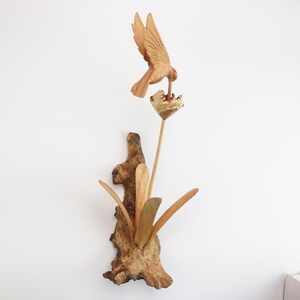 May include: A wooden sculpture of a bird perched on a branch, drinking nectar from a flower. The sculpture is made of natural wood and has a rustic, handcrafted look.