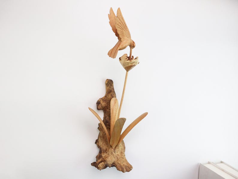 May include: A wooden sculpture of a bird perched on a branch, drinking nectar from a flower. The sculpture is made of natural wood and has a rustic, handcrafted look.