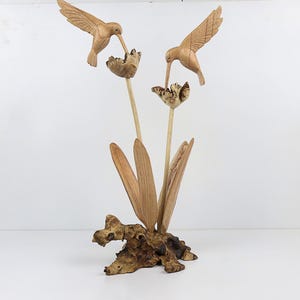 May include: A wooden sculpture of two hummingbirds perched on a branch with carved flowers. The sculpture is made of light brown wood and has a natural finish.