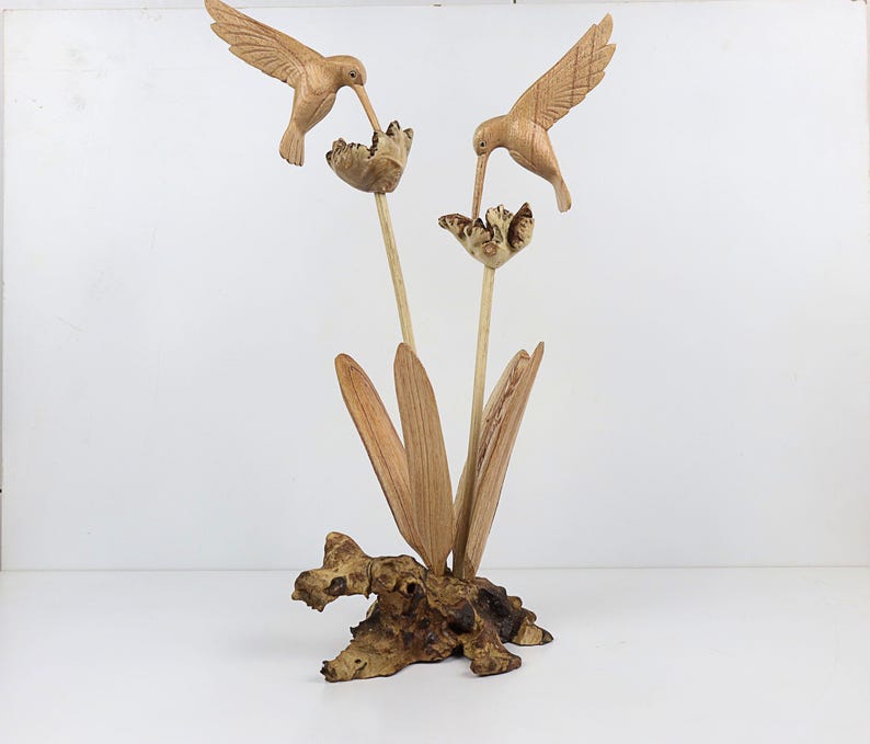 May include: A wooden sculpture of two hummingbirds perched on a branch with carved flowers. The sculpture is made of light brown wood and has a natural finish.