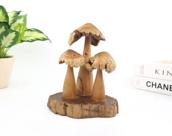 Può includere: Una scultura di funghi in legno con tre funghi di diverse dimensioni. I funghi sono marrone chiaro con cappelli e gambi strutturati, montati su una base di legno rustica. Su sfondo bianco.
