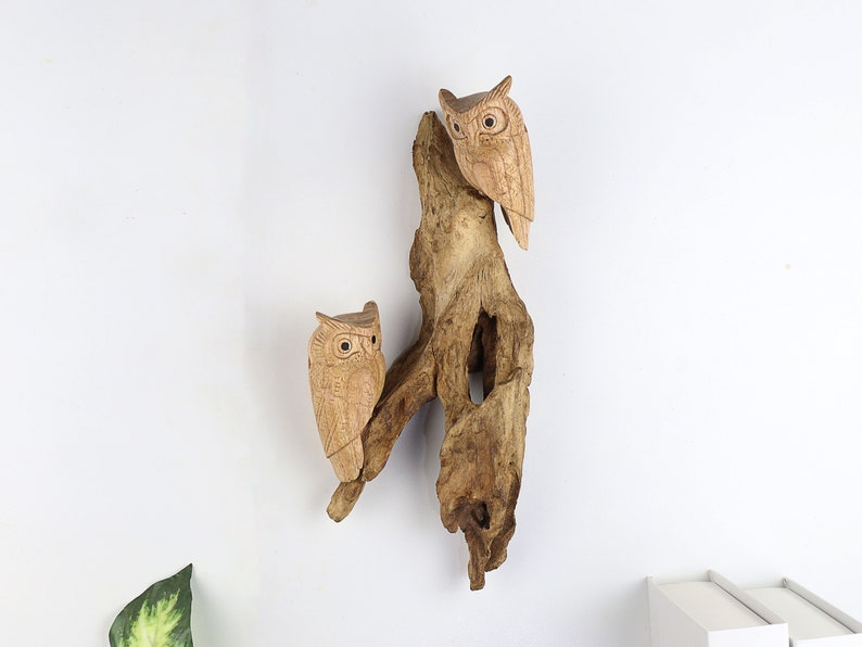 May include: Two wooden owls carved in a realistic style are perched on a piece of driftwood. The owls are brown and have large, round eyes. The driftwood is a light brown colour and has a natural, weathered look.