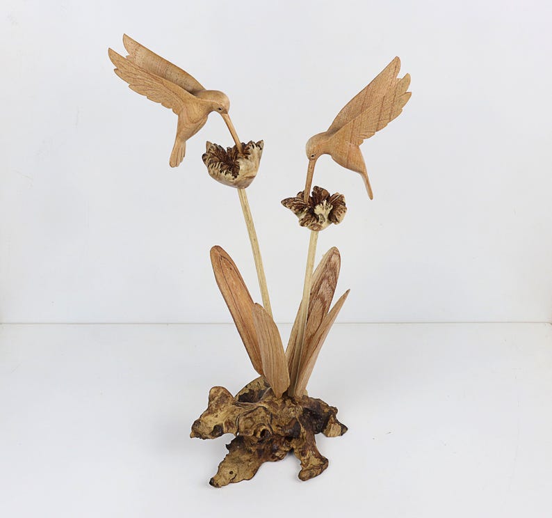 May include: A wooden sculpture of two hummingbirds perched on a branch with flowers. The sculpture is made of natural wood and has a rustic finish.