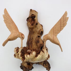 May include: A wooden sculpture of two hummingbirds perched on a branch, with a nest and eggs carved into the branch. The birds are in flight, with their wings spread out.