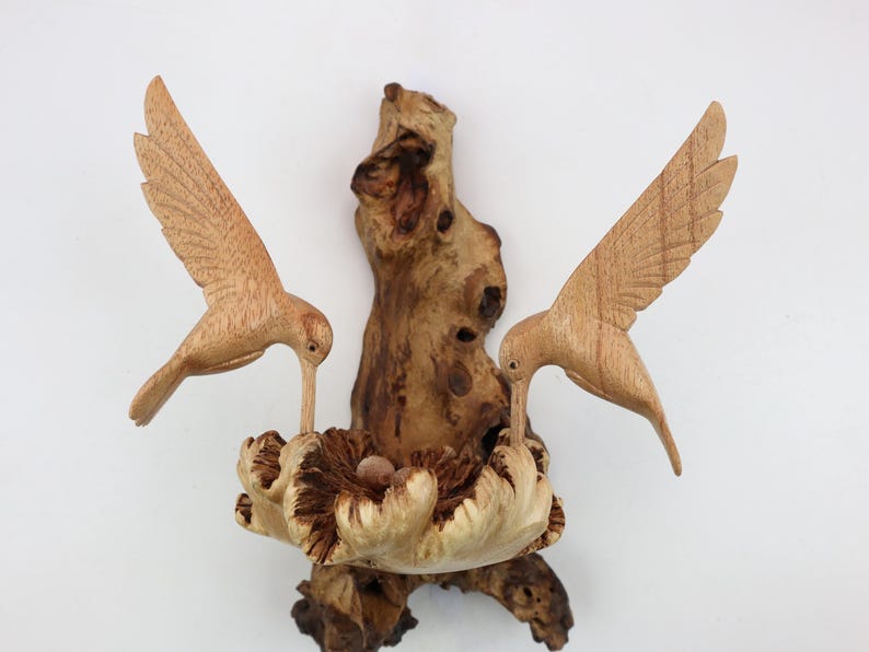 May include: A wooden sculpture of two hummingbirds perched on a branch, with a nest and eggs carved into the branch. The birds are in flight, with their wings spread out.