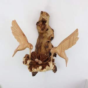 May include: Wooden wall art featuring two carved hummingbirds perched on a branch with a nest and eggs. The birds are brown and the nest is a light brown color.