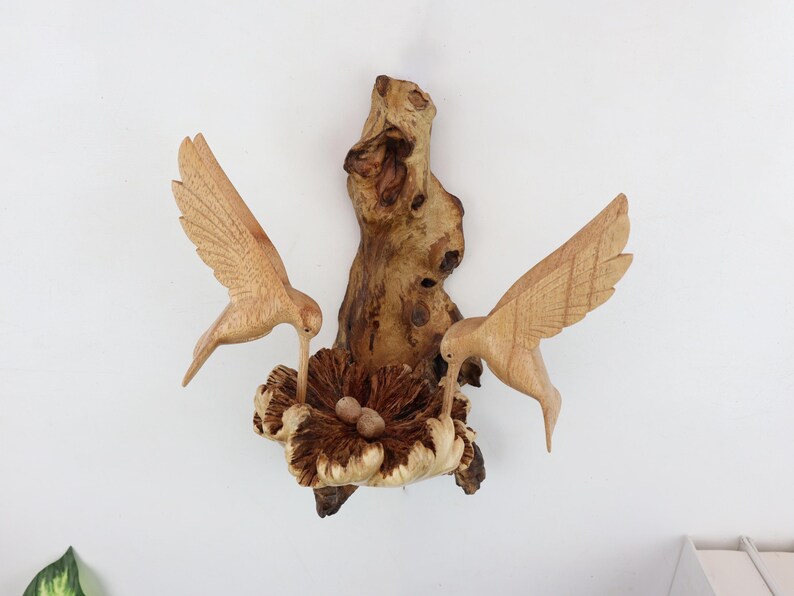 May include: Wooden wall art featuring two carved hummingbirds perched on a branch with a nest and eggs. The birds are brown and the nest is a light brown color.