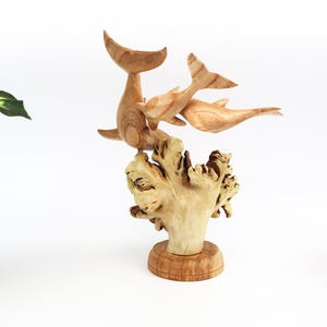 May include: A wooden sculpture of three dolphins leaping out of the water, carved in a light brown colour. The dolphins are positioned on a base that resembles a coral reef, also carved from wood.