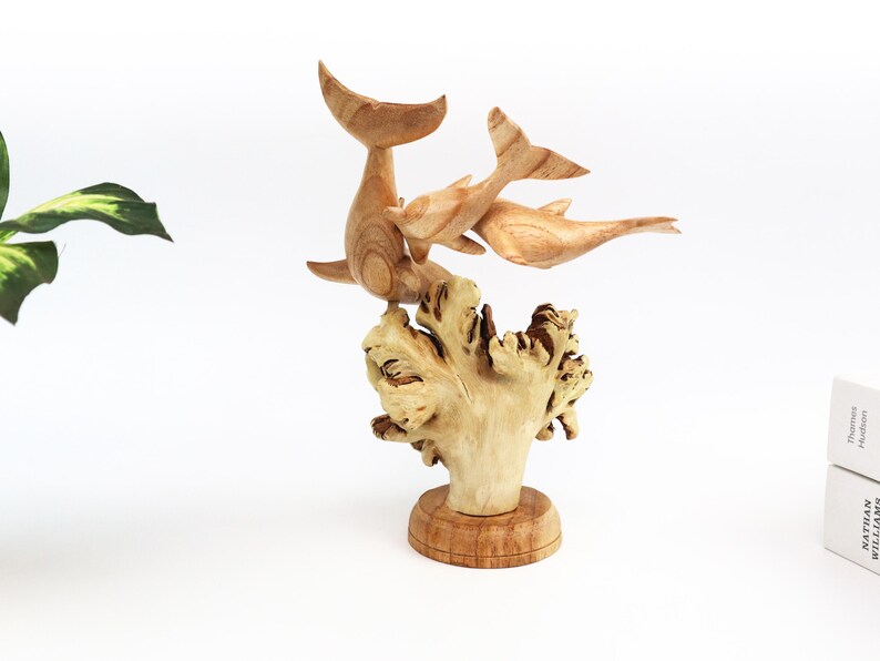 May include: A wooden sculpture of three dolphins leaping out of the water, carved in a light brown colour. The dolphins are positioned on a base that resembles a coral reef, also carved from wood.