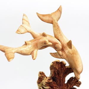 May include: A wooden sculpture of three dolphins leaping out of the water. The dolphins are carved in a realistic style and are painted in shades of brown. The sculpture is mounted on a wooden base that resembles a coral reef.