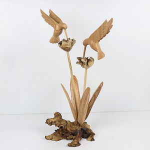 May include: Hand-carved wooden sculpture of two hummingbirds perched on a flower with leaves and a wooden base.