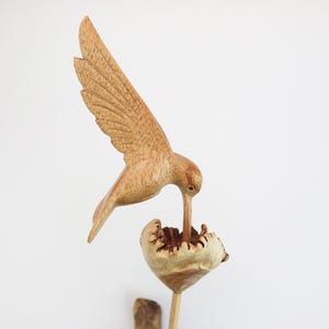 May include: A wooden hummingbird sculpture with its beak in a flower. The sculpture is made of light brown wood and has a natural finish.