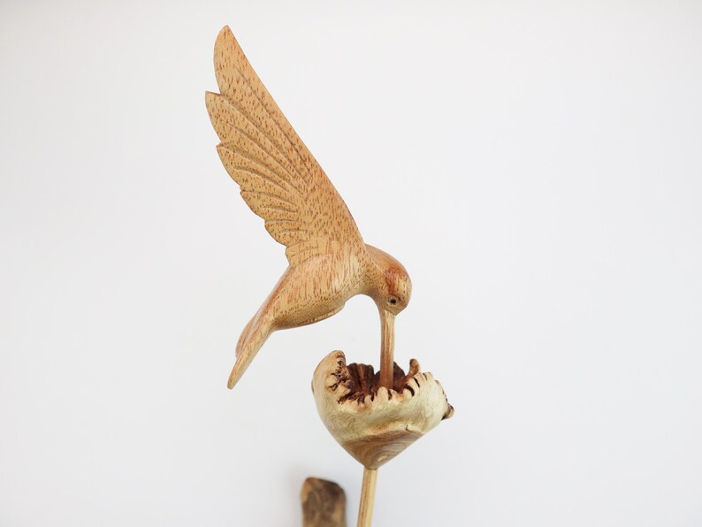 May include: A wooden hummingbird sculpture with its beak in a flower. The sculpture is made of light brown wood and has a natural finish.