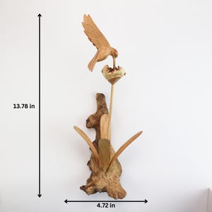 May include: A wooden sculpture of a hummingbird feeding from a flower. The sculpture is mounted on a piece of driftwood and measures 13.78 inches tall and 4.72 inches wide.
