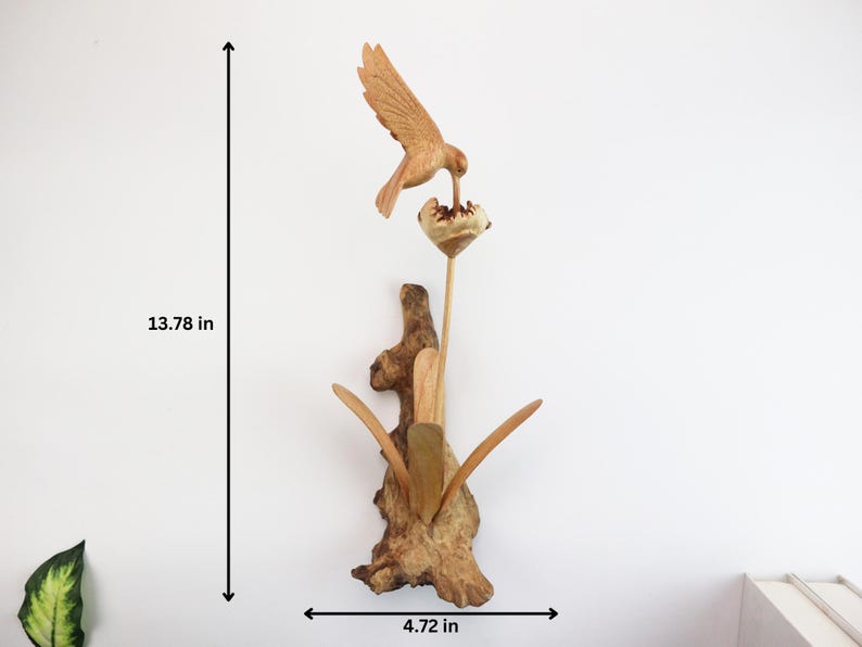 May include: A wooden sculpture of a hummingbird feeding from a flower. The sculpture is mounted on a piece of driftwood and measures 13.78 inches tall and 4.72 inches wide.