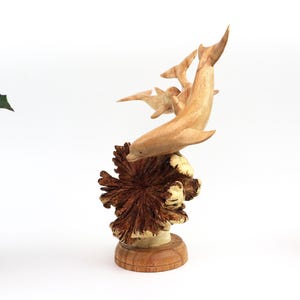 May include: Hand-carved wooden sculpture of a family of dolphins leaping out of the water. The dolphins are made of light brown wood and are set against a dark brown wooden base that resembles a coral reef.
