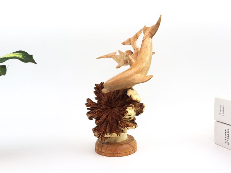 May include: Hand-carved wooden sculpture of a family of dolphins leaping out of the water. The dolphins are made of light brown wood and are set against a dark brown wooden base that resembles a coral reef.