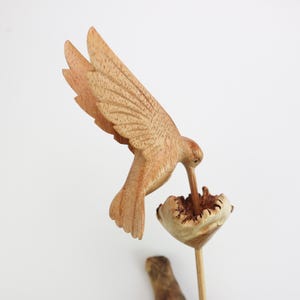 May include: A wooden hummingbird sculpture with its beak in a flower. The sculpture is painted in shades of brown and beige.