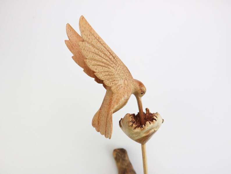 May include: A wooden hummingbird sculpture with its beak in a flower. The sculpture is painted in shades of brown and beige.