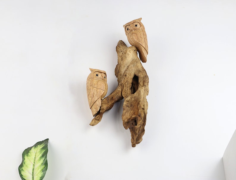 May include: Two wooden owls perched on a piece of driftwood. The owls are carved in detail and have a natural wood finish.