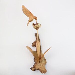 May include: A wooden sculpture of a hummingbird perched on a flower with leaves. The sculpture is made of natural wood and has a rustic, handcrafted look.