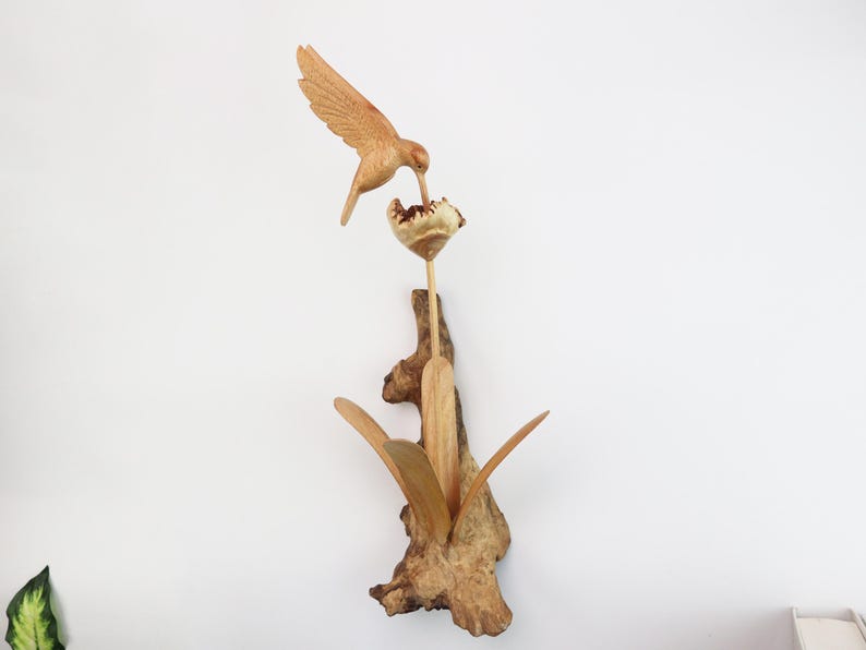 May include: A wooden sculpture of a hummingbird perched on a flower with leaves. The sculpture is made of natural wood and has a rustic, handcrafted look.