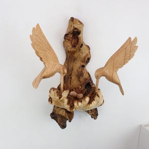 May include: A wooden sculpture of two hummingbirds perched on a branch with a nest of eggs. The sculpture is made of natural wood and has a rustic, organic look.