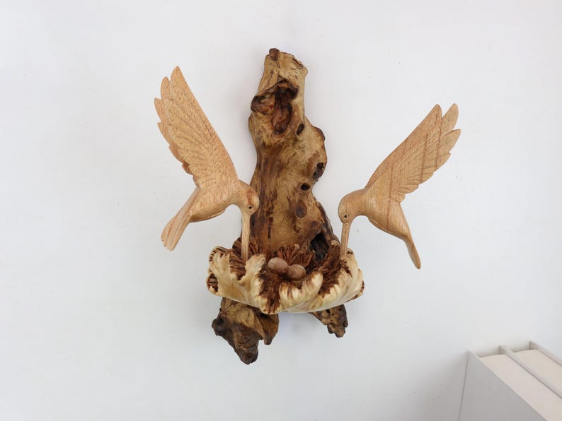 May include: A wooden sculpture of two hummingbirds perched on a branch with a nest of eggs. The sculpture is made of natural wood and has a rustic, organic look.