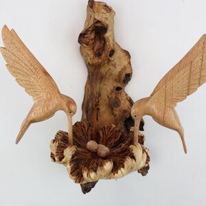May include: A wooden sculpture of two hummingbirds perched on a branch with a nest and eggs. The birds are carved in detail and the nest is made to look like a flower.