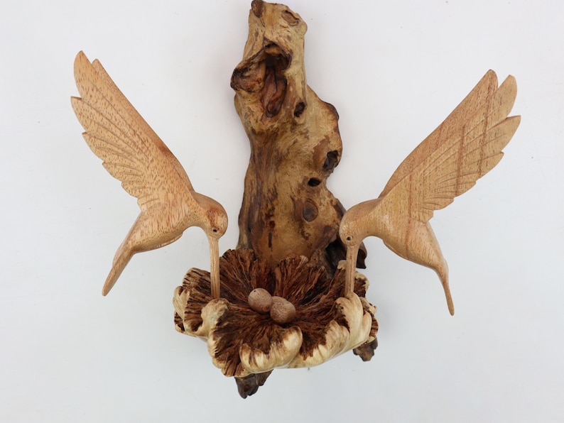 May include: A wooden sculpture of two hummingbirds perched on a branch with a nest and eggs. The birds are carved in detail and the nest is made to look like a flower.