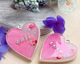 Puede incluir: Dos colgantes de resina en forma de corazón con bordes de metal plateado. El colgante izquierdo contiene pétalos de flores rosas y el texto plateado "cali". El derecho contiene pétalos rosas y el texto "cove". Ambos tienen pequeñas anillas metálicas.