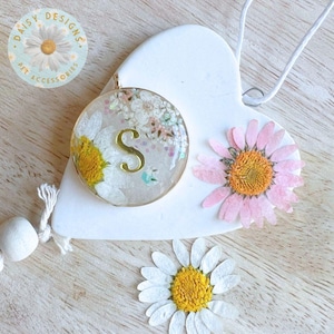 May include: A round pendant with the letter "S" in gold, encased in clear resin with dried flowers. The pendant is on a white heart-shaped base, adorned with a pink daisy and a white daisy. The logo "Daisy Designs Pet Accessories" is in the upper left.