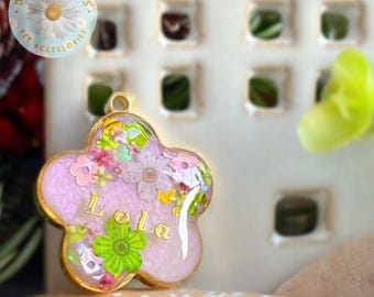 Puede incluir: Un colgante de resina con forma de flor y borde de metal dorado. El interior presenta una base de purpurina rosa claro, flores secas incrustadas en tonos rosa y verde, y el nombre Lola en letras doradas. Tiene una pequeña anilla en la parte superior.