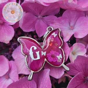 Puede incluir: Una placa para mascotas en forma de mariposa rosa con la letra "G" en plata. La placa está llena de flores secas y pequeñas mariposas blancas. La placa está rodeada de flores de hortensia rosas. La placa tiene un borde de metal plateado y un anillo para sujetar.