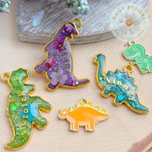 May include: Five dinosaur-shaped charms with gold-toned metal edges. Three are filled with glitter and pressed flowers, featuring the names "Night Fury," "Stitch," and "Vinner." One is a solid orange dinosaur, and one is a small green dinosaur outline.