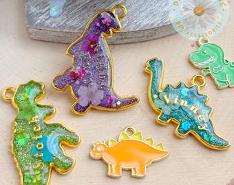 Può includere: Quattro ciondoli a forma di dinosauro con bordi in metallo dorato. Un ciondolo glitter viola con fiori pressati e il testo Night Fury, uno verde con il testo Stitch, uno blu con il testo Vinner e un ciondolo di colore arancione pieno.